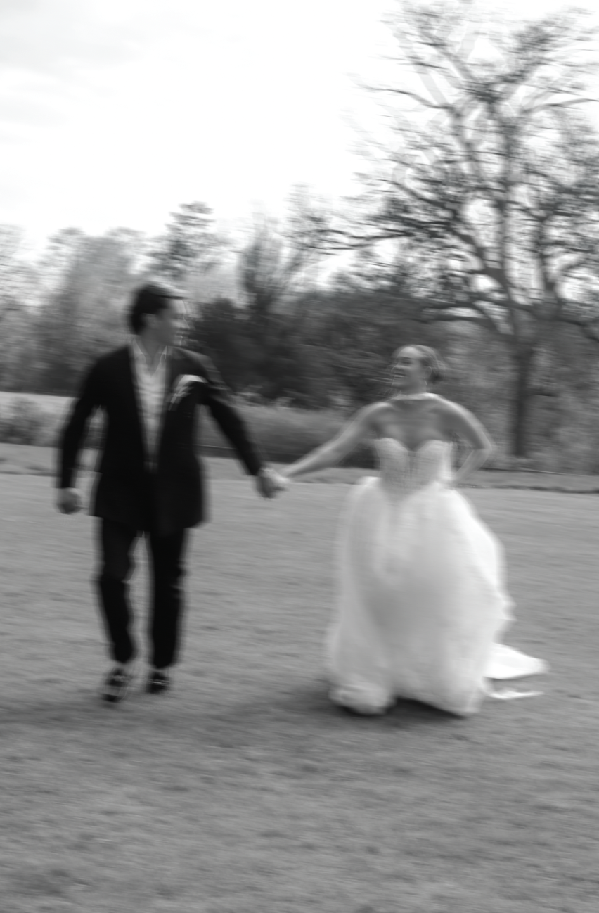 Black and white photo of a bride and groom running hand in hand across a lawn